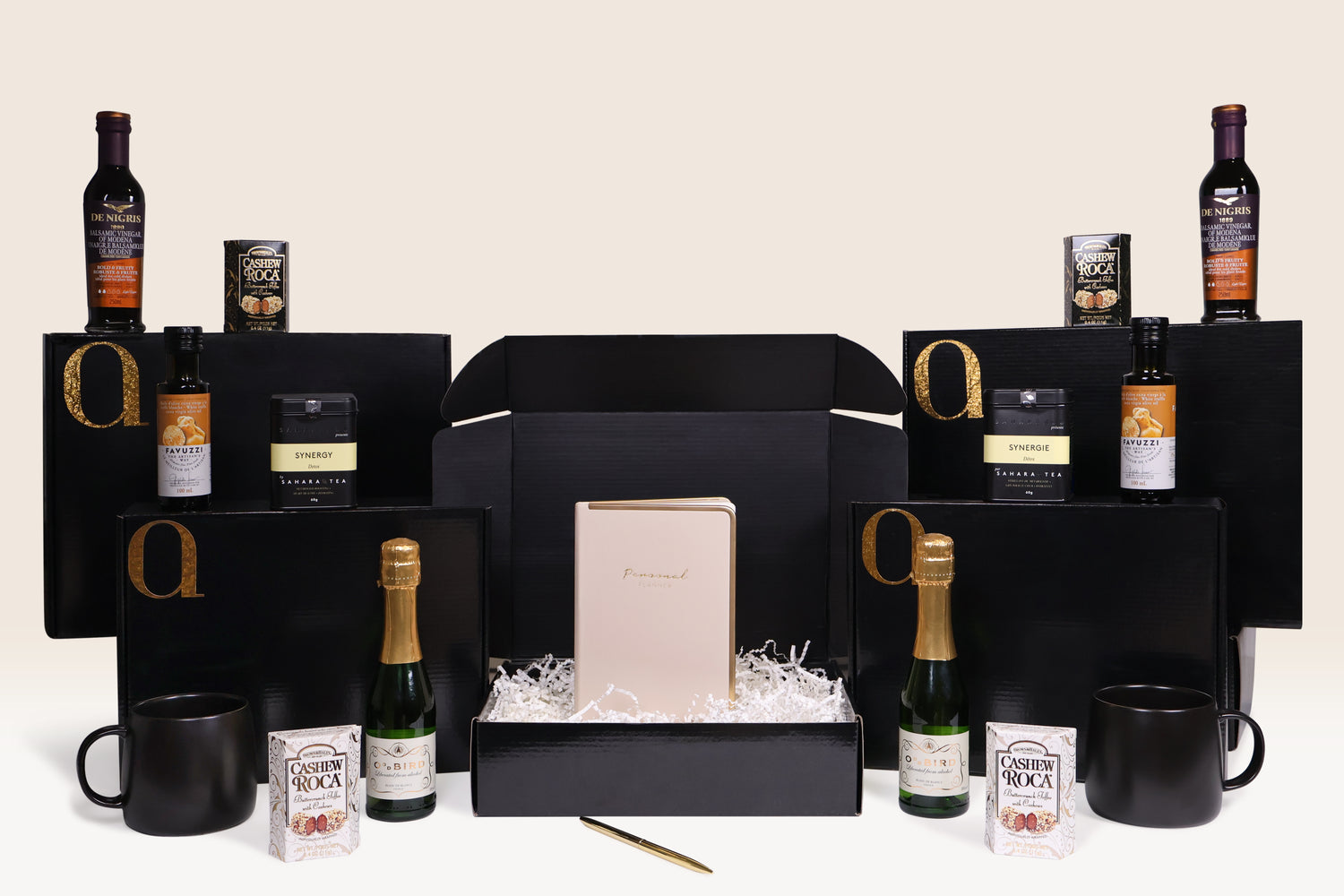 An elegant gift box arrangement featuring gourmet items, a notebook, tea, beverages, and chocolates on a neutral background.