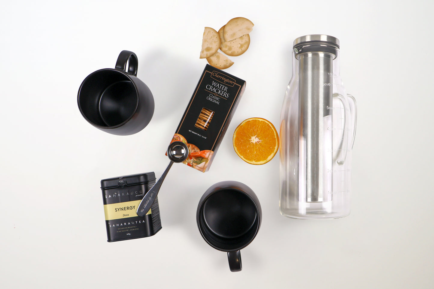 Curated tea gift box items including black mugs, crackers, loose-leaf tea, glass tea infuser, measuring spoon, and an orange slice on white background.