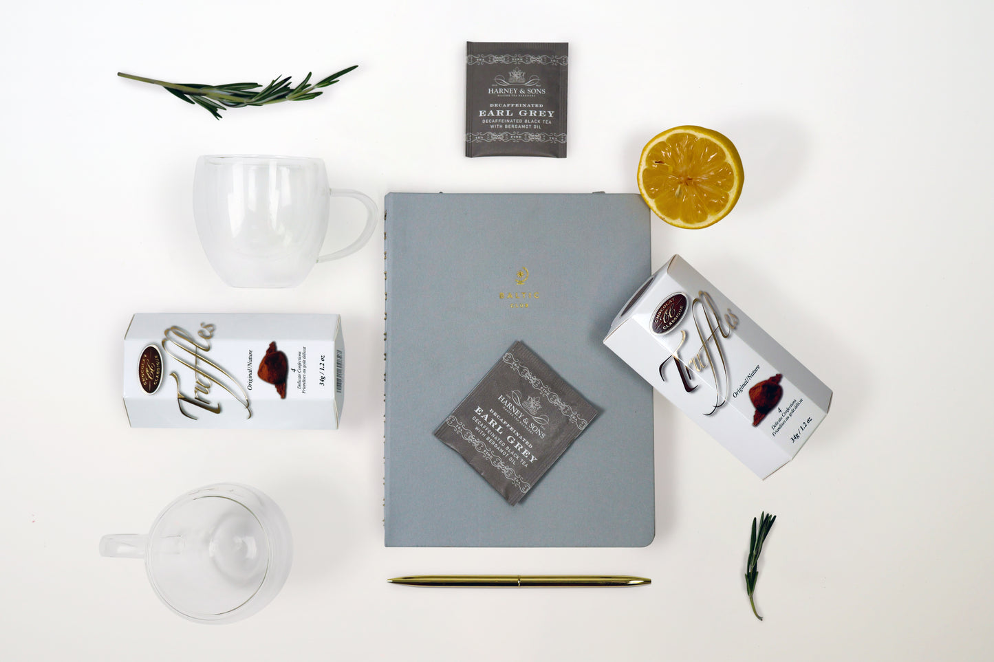 Flat lay of productivity gift box items including a grey planner, Harney & Sons tea, Belgian truffles, gold pen, double wall glass mugs, and greenery on a white background.