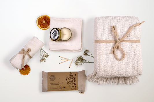Flat lay of Relaxation Retreat gift box items including a woven throw blanket, lavender eye pillow, soy candle, wash cloths, and dried citrus.